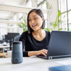 Asian businesswoman talking to virtual assistant at her desk. Female professional working on laptop and talking into a speaker.